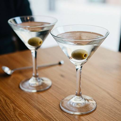 Photograph of two martini glasses with clear liquid and green olives on a wooden table, blurry background, spoon to the left.