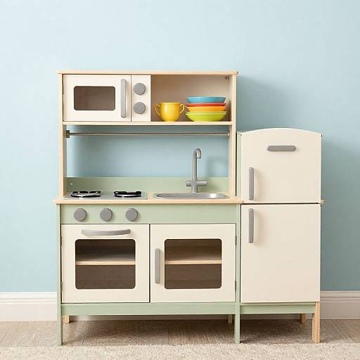 Photograph of a vintage-style, cream-colored wooden play kitchen with microwave, stove, sink, fridge, and shelves holding colorful bowls, set against a