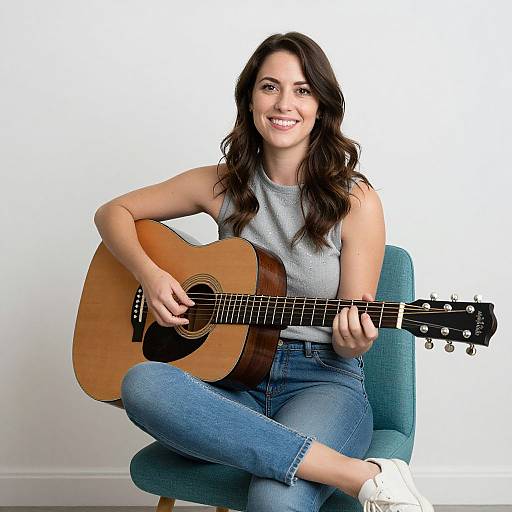 Photograph of a smiling woman with long brown hair, wearing a sleeveless gray top and blue jeans, playing an acoustic guitar while seated on a teal