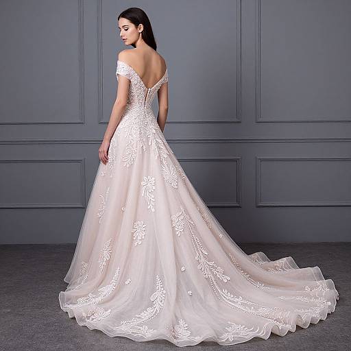 Photograph of a woman in an off-the-shoulder, white lace wedding gown with a long, flowing train, standing against a gray paneled