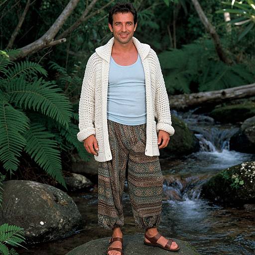 Photograph of a smiling man with short black hair, wearing a white crochet cardigan, light blue tank top, patterned pants, and brown sandals