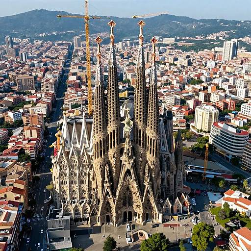 Aerial photograph of Barcelona's Sagrada Família, showcasing its intricate Gothic spires amidst a densely packed urban landscape with mountains in the background. Bright