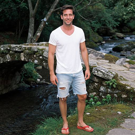 Photograph of a smiling, bearded man in a white V-neck T-shirt, ripped light blue shorts, and red flip-flops, standing by