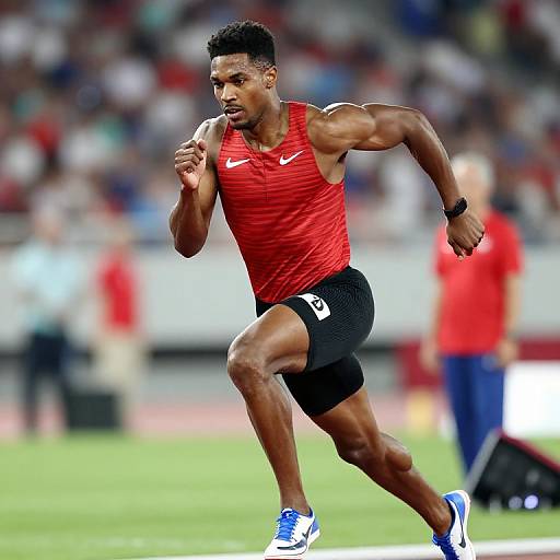 Photograph of a muscular, black male sprinter in a red sleeveless shirt and black shorts, running on a track, with a blurred crowd in