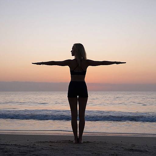Silhouetted woman with outstretched arms stands on sandy beach at sunset, waves gently in background, sky transitions from pink to blue.