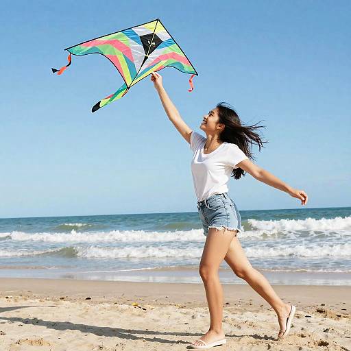 Young woman in white t-shirt and denim shorts flying a colorful kite on a sunny beach with clear blue sky and waves.