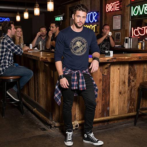Photograph of a bearded man in a black long-sleeve shirt, plaid shirt tied around waist, black jeans, and Converse shoes