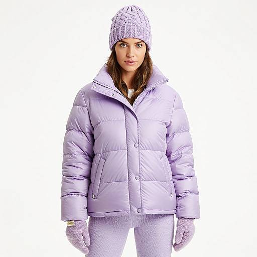 Photograph of a woman with light skin, brown hair, wearing a white knit hat, light purple puffer jacket, white gloves, and white pants