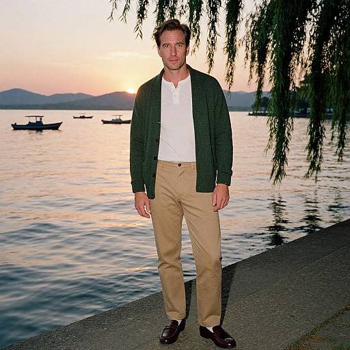 Photograph of a man with short brown hair, wearing a green cardigan, white shirt, beige pants, and brown shoes, standing on a river