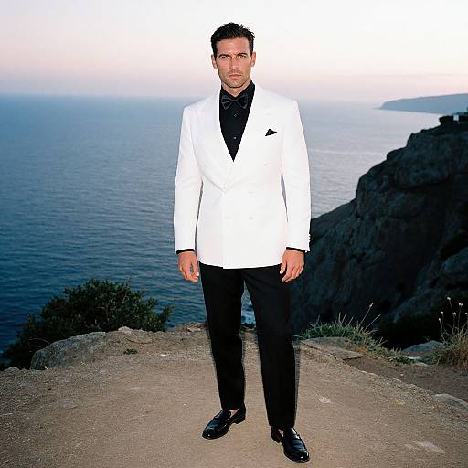 Photograph of a man with short dark hair, wearing a white suit, black shirt, and black shoes, standing on a rocky cliff overlooking a vast