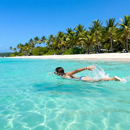 Photograph of a swimmer with dark hair, wearing black swim trunks, swimming in clear turquoise water near a tropical beach with palm trees and a