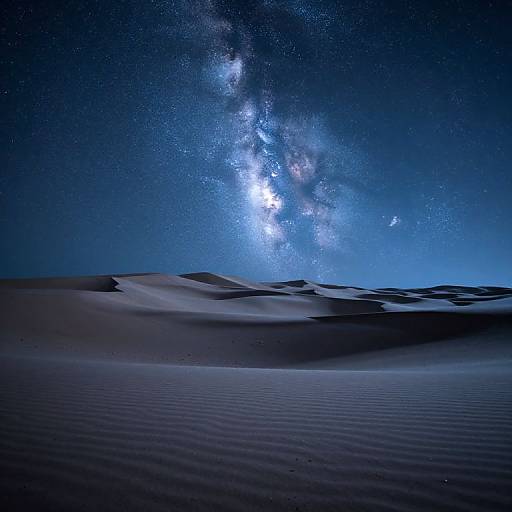 Photograph of a starry night sky over a serene desert with rippled sand dunes, displaying the Milky Way galaxy in vibrant blue and white.