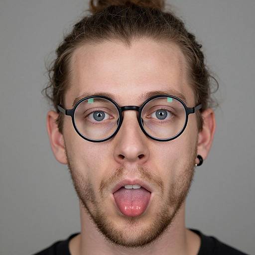Photograph of a man with blue eyes, round black glasses, brown hair in a bun, beard, black earring, and tongue out against a
