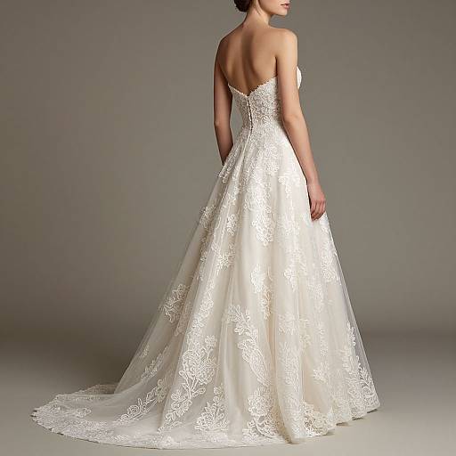 Photograph of a woman in an elegant white lace wedding gown with a strapless back and a full, flowing train, standing against a gray studio background