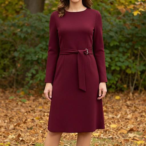 Photograph of a woman in a burgundy long-sleeve dress with a tied belt, standing in a forest with autumn leaves.