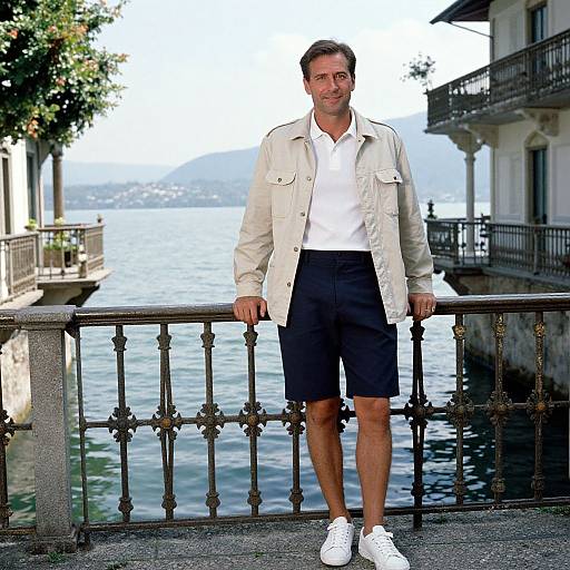 Photograph of a man with short brown hair, wearing a white shirt, beige jacket, black shorts, and white sneakers, standing by a waterfront railing