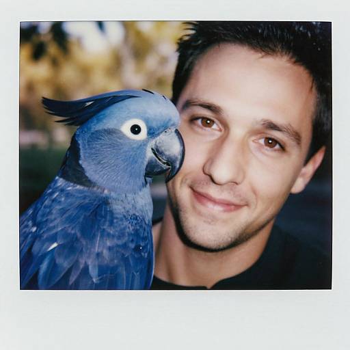Photograph of a smiling young man with short dark hair and fair skin, closely positioned next to a blue parrot with a black beak. Bl