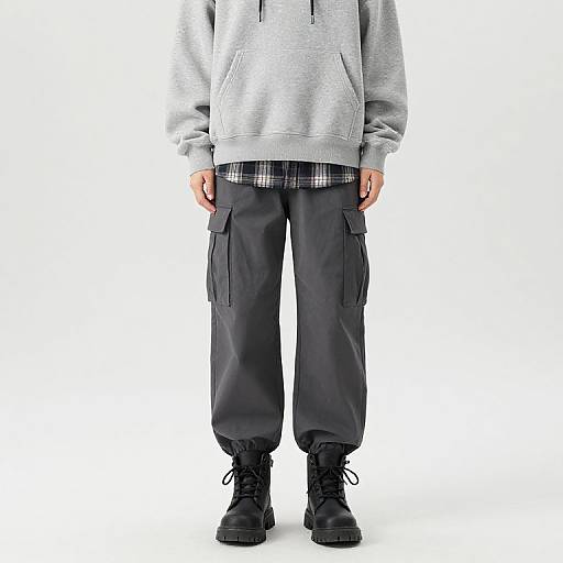 Photograph of a person wearing a gray hoodie, black cargo pants, black boots, and a plaid shirt underneath, standing against a white background.