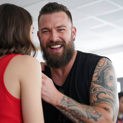 Photograph of a smiling bearded tattooed man in black sleeveless shirt, talking to a woman in red sleeveless top, bright background.