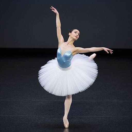 Photograph of a graceful ballerina in a blue bodice and white tutu, balancing on pointe, arms elegantly extended against a dark