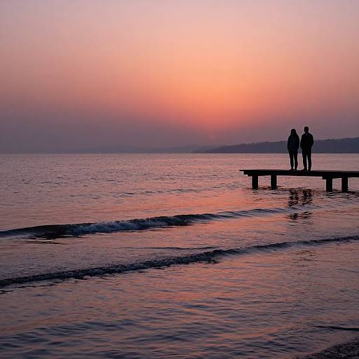 Silhouetted couple stands on pier at sunset, reflecting pink and orange sky over calm, rippling water. Photographic image.