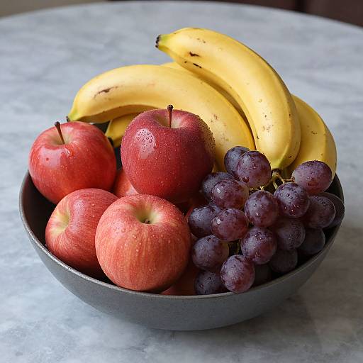 Photograph of a bowl containing red apples, ripe bananas, and purple grapes, all with water droplets, on a marble countertop.