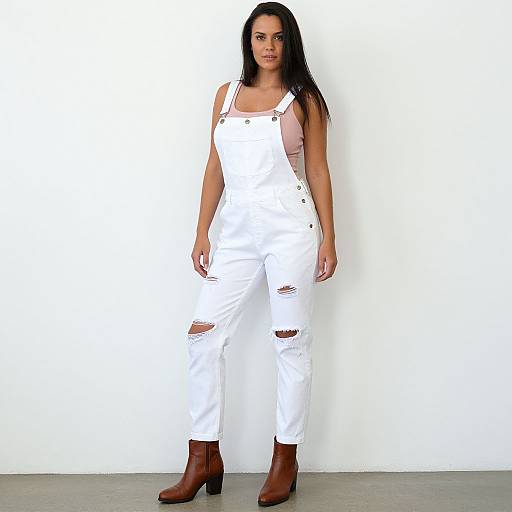 Photograph of a confident woman with long black hair, wearing white ripped overalls, pink tank top, brown boots, standing against a plain white wall