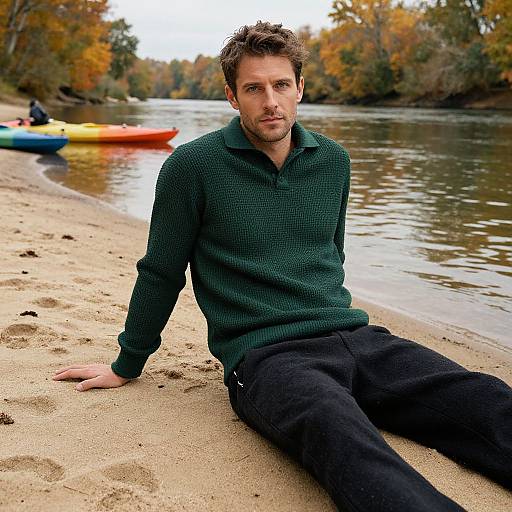 Photograph of a handsome, brown-haired man in a green sweater and black pants, sitting on a sandy riverbank with colorful kayaks in the background