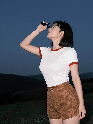 Photograph of a light-skinned woman with black hair, wearing a white shirt with red trim and brown shorts, looking through binoculars at twilight