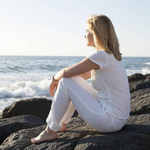 Photograph of a blonde woman in white clothing, sitting on black rocks by the ocean, facing the sunlit sea, barefoot, hands resting on