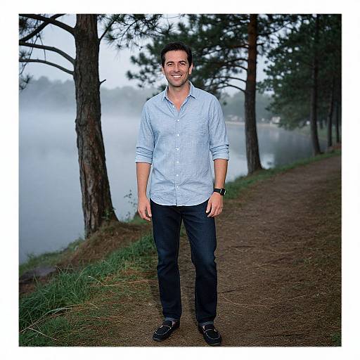 Photograph of a smiling man with short dark hair, wearing a light blue button-up shirt, black pants, and black shoes, standing on a dirt