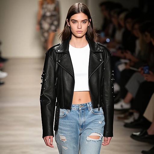 Photograph of a brunette model with straight hair, wearing a black leather jacket, white crop top, and distressed blue jeans, walking down a brightly lit