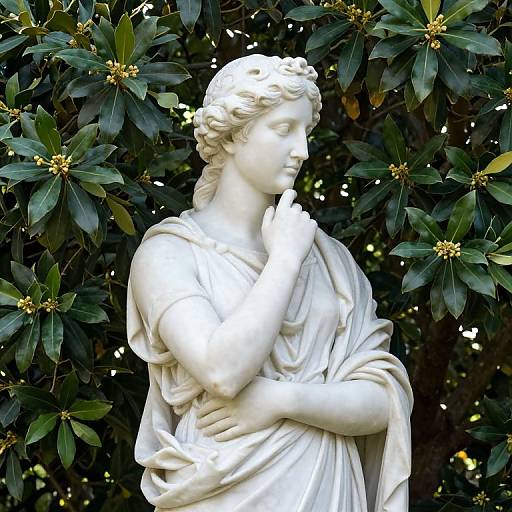 Photograph of a white marble statue of a serene, classical woman with wavy hair, draped in flowing robes, standing against lush, dark green foliage