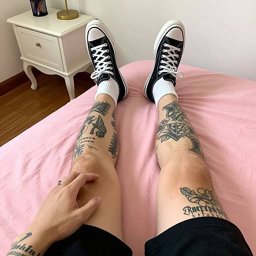 Photograph of a tattooed person's legs wearing black Converse sneakers and white socks, lying on a pink bed, with a hand on their thigh