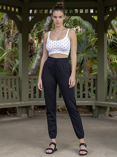 Photograph of a slim woman with dark hair in a bun, wearing a white polka dot crop top and black high-waisted pants, standing