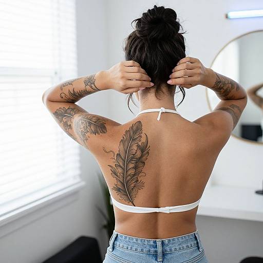Photograph of a tattooed woman with dark hair in a bun, adjusting her white bikini top, showing a large feather tattoo on her back, standing