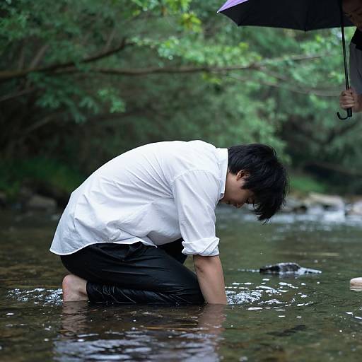 Photograph of a man with wet black hair, white shirt, and black pants, kneeling in a shallow, rain-soaked forest stream, holding an