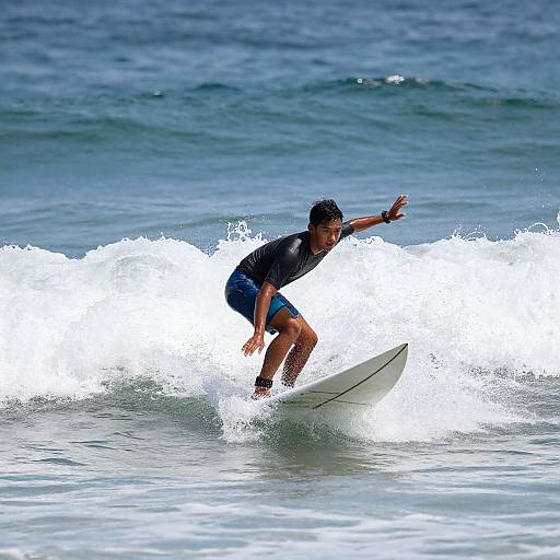 Photograph of a young male surfer with dark hair, wearing a black wetsuit top and blue shorts, riding a white-capped wave in