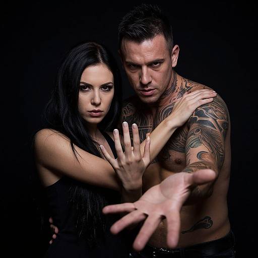 Photograph of a tattooed, muscular man with short black hair and a pale-skinned, dark-haired woman with intense expressions, reaching forward against a