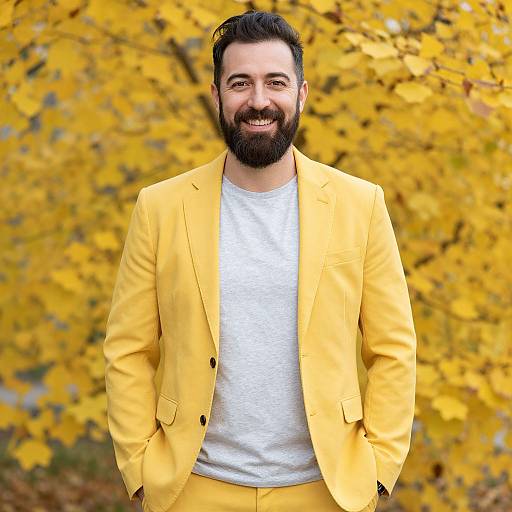 Photograph of a smiling bearded man with dark hair, wearing a yellow blazer and white t-shirt, standing against a vibrant yellow autumn leaf background