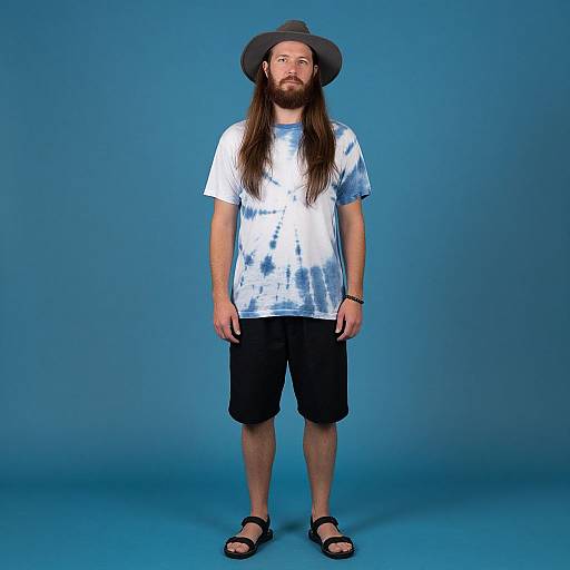 Photograph of a bearded man with long brown hair, wearing a gray hat, white blue tie-dye shirt, black shorts, black sandals,
