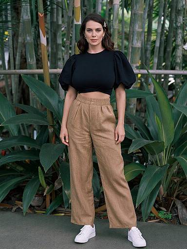 Photograph of a woman with long dark hair, wearing a black crop top, high-waisted tan pants, and white sneakers, standing in a