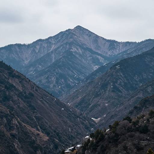 Photograph of a mountainous landscape with dark green and brown hills, snow patches, and a large, misty gray peak in the background under a