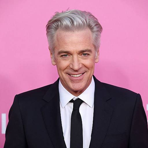 Photograph of an elderly man with silver hair, smiling, wearing a black suit, white shirt, and black tie, against a bright pink background.