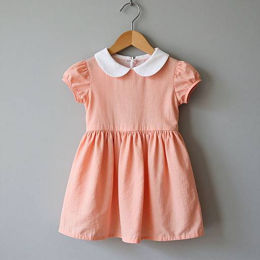 Photograph of a peach-colored, short-sleeved, white-collared children's dress, hanging on a wooden hanger against a gray wall