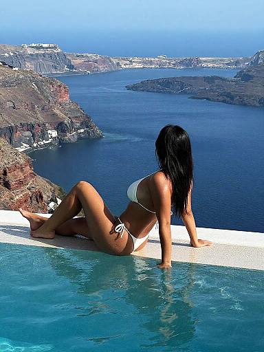 Photograph of a woman with long black hair in a white bikini, lounging at the edge of a pool overlooking a stunning blue sea and mountainous