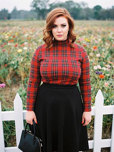 Photograph of a curvy, fair-skinned woman with wavy auburn hair, wearing a red plaid turtleneck and black skirt
