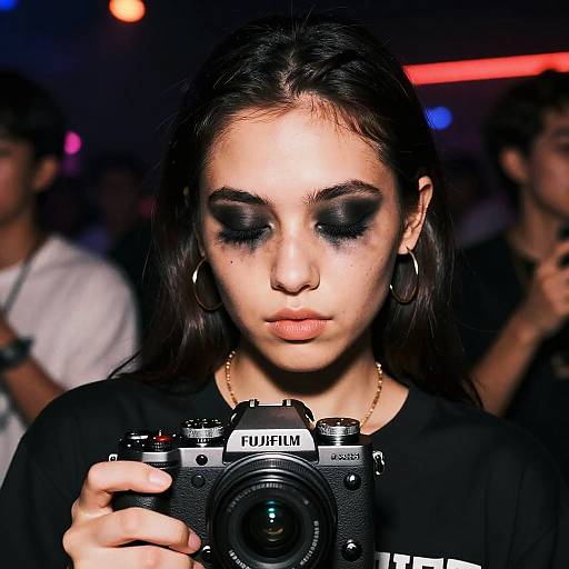 Photograph of a young woman with dark hair, heavy black eyeshadow, wearing hoop earrings and a black shirt, holding a Fujifilm camera