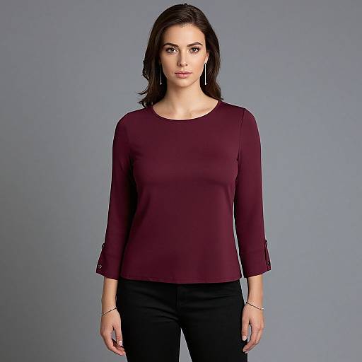 Photograph of a young woman with medium-length brown hair, wearing a maroon long-sleeve top and black pants, standing against a plain gray