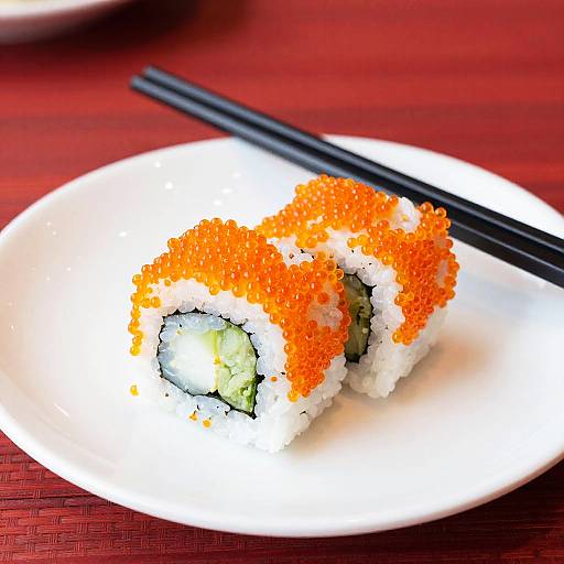 Photograph of two sushi rolls with bright orange tobiko on a white plate, black chopsticks on a red woven tablecloth.